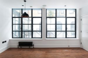 A bright, modern room with large steel-look windows featuring a black grid design, offering a view of an urban landscape, complemented by a classic dark metal radiator and a wooden floor.
